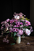 Bouquet of fresh colorful peonies and chrysanthemums in white vase placed on wooden table in dark room