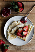 Top view of delicious crepes with sweet strawberry jam placed on plate near spoon on wooden table