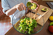 Crop woman in casual clothes putting avocado cubes into glass bowl with lettuce while cooking vegan salad for lunch in sunlit kitchen