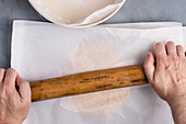 From above of crop anonymous female preparing dough covered with baking paper with wooden rolling pin on table while preparing cake