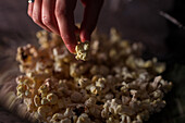 Abgeschnittene, nicht erkennbare Frau in Freizeitkleidung sitzt mit gekreuzten Beinen auf der Couch und isst Popcorn zu Hause