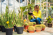 Female gardener using modern tablet while counting plants and working in garden center