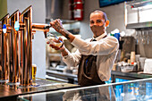 Fröhlicher männlicher Barkeeper in Uniform und Schutzmaske füllt ein Glas mit Bier aus dem Zapfhahn, während er am Tresen einer Kneipe während der Coronavirus-Pandemie arbeitet