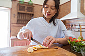 Young Asian female cutting fresh banana on chopping board near bowl with salad while cooking healthy food in sunlit kitchen