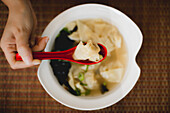 Hand von oben, die eine Schüssel mit leckerer heißer Wonton-Suppe mit rotem Löffel in einem asiatischen Restaurant isst