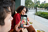 Crop-Mann mit Smartphone fotografiert fröhliche ethnische Freundin beim gesunden Frühstück mit Saft und Salat am Tresen eines modernen Restaurants