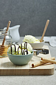 Leckere Eiskugeln in einer Schale mit Schokoladensoße, serviert auf einem hölzernen Schneidebrett mit einem Glas Honig und Waffelröllchen in der Küche