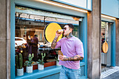 Young cool male in sunglasses enjoying takeaway hot drink while walking on street with tasty sandwich in wrapper