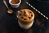 Von oben ein Glas süßer Dalgona-Kaffee mit schaumigem Belag, serviert auf einem Tisch mit Schokoladenwaffelrolle und Kakaopulver