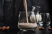 Pouring fresh aromatic cinnamon milkshake into glass placed on black table near almonds and sticks