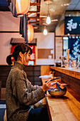 Side view of pleased Asian woman in casual wear laughing while sitting at counter with chopsticks and bowl with ramen in cafe