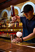 Young Asian bartender pouring strawberry juice in the shaker while preparing a cocktail in the bar