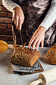 Anonyme Bäckerin mit Messer schneidet frisches Brot mit Sonnenblumenkernen auf einem Tisch in einer Backstube