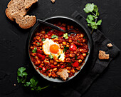 Top view of chakchouka with sunny side up egg in delicious tomato sauce with rye bread piece in bowl between fresh cilantro on dark background