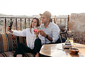 Positives, junges, stilvolles Paar, das auf einer Couch sitzt und mit einem Cocktail anstößt, während es ein Selbstporträt mit dem Handy auf einer Terrasse in Kappadokien, Türkei, macht