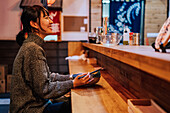 Seitenansicht einer zufriedenen asiatischen Frau in Freizeitkleidung, die lachend am Tresen mit Stäbchen und Schüssel mit Ramen in einem Café sitzt