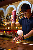 Young Asian bartender pouring strawberry juice in the shaker while preparing a cocktail in the bar