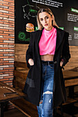 Attractive young female in jeans pink sweater and black coat standing with hands in pockets near table in restaurant and looking at camera