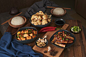 Mala-Tofu und Yuxiang, chinesische vegane Gerichte, begleitet von einer Schüssel mit Reis, Blumenkohl, Sojasauce und einer japanischen Teekanne auf einem mit Stoffen dekorierten Holztisch