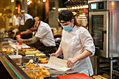 Through glass of female chef wearing white jacket checking paper sheets while standing at counter in kitchen with busy cooks preparing food during coronavirus pandemic