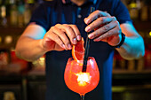 Bildausschnitt mit den Händen eines jungen Barkeepers, der einen Cocktail mit Grapefruitkeil und Strohhalmen in einer Bar dekoriert
