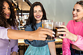 Joyful young multiethnic female best friends in stylish colorful outfits smiling and clinking glasses of refreshing juice and coke in modern bar