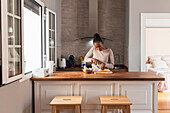 Schwarze Frau in Kleidung mit gestreiftem Ornament schneidet Kochbanane mit Messer auf Schneidebrett an Tisch in Haus