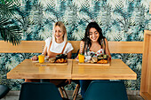 Young content best female friends sitting at wooden table with tasty croissants and beverages in cafeteria