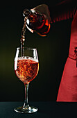 Crop unrecognizable barman in red apron pouring semi sweet fine red wine into wineglass in dark studio
