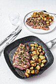 From above of appetizing fried beef chop with potatoes topped with minced garlic and parsley served on frying pan and plate on white background with cutlery