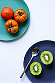 Draufsicht auf frische, gesunde halbierte Kiwi und ganze Tomaten auf blauen und türkisfarbenen runden Tellern mit Löffel auf weißem Hintergrund