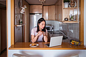 Fokussierte Asiatin mit heißem Getränk surft auf ihrem Netbook, während sie beim Frühstück in einer modernen Küche zu Hause am Tresen sitzt