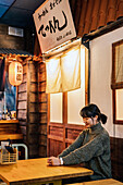 Zufriedene asiatische Frau in legerem Pullover, die an einem Holztisch in einer Ramen-Bar sitzt und nach unten schaut
