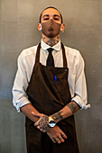 Männlicher Kellner mit Tattoos in weißem Hemd und Schürze schaut in die Kamera, während er auf grauem Hintergrund in einem modernen Café arbeitet