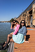 Cheerful multiethnic female friends in casual outfits sitting on city waterfront and looking at camera with smiles on sunny summer day
