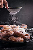 Crop anonymous chef with strainer sprinkling powdered sugar on heap of sweet fried doughnuts served on plate on gray background