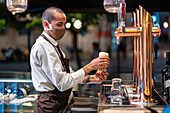 Männlicher Barkeeper in Uniform und Schutzmaske füllt ein Glas mit Bier aus dem Zapfhahn, während er am Tresen in einer Kneipe während einer Coronavirus-Pandemie arbeitet