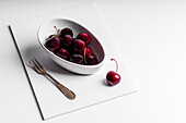 High angle of healthy ripe tasty cherries in ceramic bowl placed with fork on white board