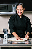 Young smiling ethnic female chef in uniform taking video on smartphone while cooking delicious pizza on kitchen counter