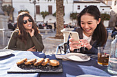 Smiling Asian female taking picture of tasty fired snack on cellphone near female friend while sitting at table in outdoor cafeteria