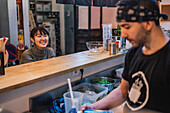 Asian woman in casual wear sitting at counter and talking with male worker of modern ramen bar
