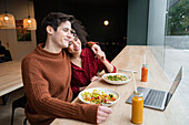 Fröhliches multiethnisches Paar isst gesundes Frühstück im Restaurant und surft auf dem Laptop