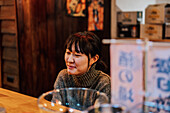 Asiatische Frau in Freizeitkleidung sitzt am Holztresen und wartet auf ihre Bestellung in einer Ramen-Bar