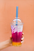 Hand of anonymous female holding takeaway cup of tasty ice juice against orange wall on street