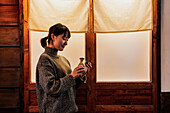 Seitenansicht einer überglücklichen Asiatin im Pullover, die mit einem traditionellen Getränk in einem Café an der Tür steht