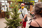 Älterer männlicher Einkäufer in steriler Maske mit Nadelbaum im Einkaufswagen, der gegen eine unerkennbare Frau in einem Gartengeschäft blickt