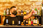 Blurred barman in sterile mask standing at bar counter and cleaning glass while working in restaurant during coronavirus