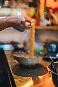 Hand eines gesichtslosen Kochs mit hölzernen Essstäbchen, der eine Nudel über einer Schüssel auf einem unscharfen Hintergrund in einer Ramen-Bar hält
