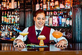 Smiling female barkeeper standing at bar counter with different types of alcohol drinks served in creative cocktail glasses in shape of mushroom and fish