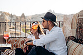 Seitenansicht eines jungen bärtigen Mannes mit Sonnenbrille und modischem Hut, der auf einer Terrasse in Kappadokien, Türkei, einen kalten erfrischenden Cocktail schlürft